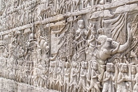 An intricate stone bas-relief depicts a historical scene with numerous figures in an ancient style. The scene includes soldiers, elephants, and chariots, suggesting a procession or battle. The detailed carvings cover the surface, showing various human figures and animals engaged in dynamic interactions.