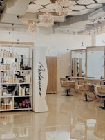A modern hair salon with elegant design elements. The space features built-in shelves stocked with hair products, large mirrors, and cushioned salon chairs. The ceiling is decorated with geometric patterns and several spherical light fixtures. A mural of abstract line art adorns the walls.