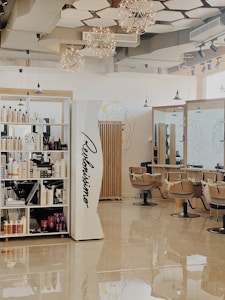 A modern hair salon with elegant design elements. The space features built-in shelves stocked with hair products, large mirrors, and cushioned salon chairs. The ceiling is decorated with geometric patterns and several spherical light fixtures. A mural of abstract line art adorns the walls.