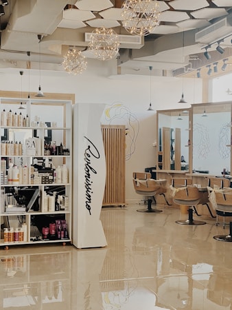 A modern hair salon with elegant design elements. The space features built-in shelves stocked with hair products, large mirrors, and cushioned salon chairs. The ceiling is decorated with geometric patterns and several spherical light fixtures. A mural of abstract line art adorns the walls.