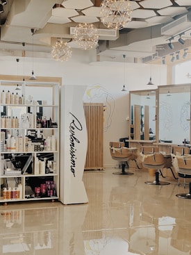 A modern hair salon with elegant design elements. The space features built-in shelves stocked with hair products, large mirrors, and cushioned salon chairs. The ceiling is decorated with geometric patterns and several spherical light fixtures. A mural of abstract line art adorns the walls.