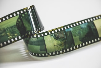 A roll of photographic film features several frames of urban scenes. The film has a prominent green tint and is coiled slightly, resting on a light surface. The film edges feature sprocket holes and some text indicating 'AGFA EXCL CTX 100'.