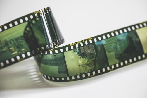 A roll of photographic film features several frames of urban scenes. The film has a prominent green tint and is coiled slightly, resting on a light surface. The film edges feature sprocket holes and some text indicating 'AGFA EXCL CTX 100'.
