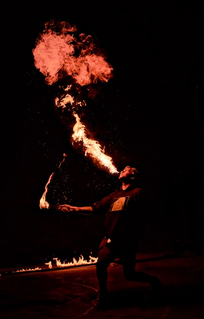 A person is performing fire breathing against a dark background. The fire creates a dramatic, bright orange flame that contrasts with the darkness. The performer appears to be in motion, with one hand holding a torch and their face illuminated by the flames.