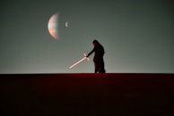 A silhouetted figure stands on a dark horizon wielding a glowing red lightsaber. The background features a large planet and its moon set against a dark sky.