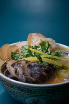 A bowl of soup with rich ingredients including a large mushroom, sliced vegetables, and fresh herbs. The bowl is ornately decorated with a traditional pattern and the background is a plain teal color, enhancing the focus on the dish.