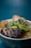 A bowl of soup with rich ingredients including a large mushroom, sliced vegetables, and fresh herbs. The bowl is ornately decorated with a traditional pattern and the background is a plain teal color, enhancing the focus on the dish.