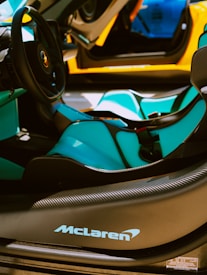 The interior of a luxury sports car is featured, showcasing sleek black and turquoise seats, a stylish steering wheel, and prominent branding on the dashboard. The car's door is open, revealing an elegant design with a vibrant orange exterior.