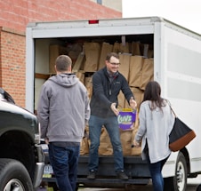 People are unloading items from the back of a white truck. The truck is filled with brown paper bags, and one person is handing a package of diapers to another individual.
