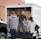 People are unloading items from the back of a white truck. The truck is filled with brown paper bags, and one person is handing a package of diapers to another individual.