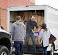 People are unloading items from the back of a white truck. The truck is filled with brown paper bags, and one person is handing a package of diapers to another individual.