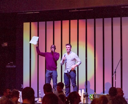Two people are standing on a stage, each holding a microphone. One person is holding a piece of paper aloft. The background has vertical light panels with a warm, colorful gradient. They appear to be addressing an audience in a dimly lit room, with several people visible in the foreground.