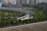A curved elevated roadway stretches through an area with lush greenery, bordered by neatly arranged trees and surrounded by a cityscape of tall residential buildings. The road is lined with safety barriers, and streetlights are visible along the route. The environment combines urban and natural elements.