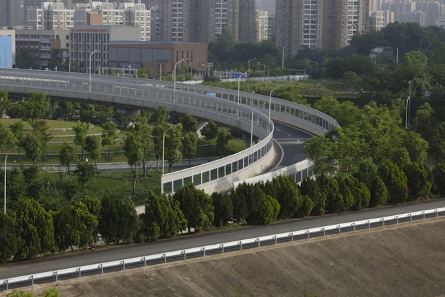 A curved elevated roadway stretches through an area with lush greenery, bordered by neatly arranged trees and surrounded by a cityscape of tall residential buildings. The road is lined with safety barriers, and streetlights are visible along the route. The environment combines urban and natural elements.