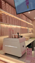 A brightly-lit interior with a pink and white color scheme features a wall of stacked pink pizza boxes. A white and pink coffee machine labeled 'Radiofonica' sits on a counter, accompanied by branded pink and white cups, napkins, and dispensers. A digital screen above displays an ice cream cone graphic.