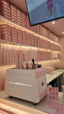 A brightly-lit interior with a pink and white color scheme features a wall of stacked pink pizza boxes. A white and pink coffee machine labeled 'Radiofonica' sits on a counter, accompanied by branded pink and white cups, napkins, and dispensers. A digital screen above displays an ice cream cone graphic.