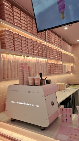 A brightly-lit interior with a pink and white color scheme features a wall of stacked pink pizza boxes. A white and pink coffee machine labeled 'Radiofonica' sits on a counter, accompanied by branded pink and white cups, napkins, and dispensers. A digital screen above displays an ice cream cone graphic.