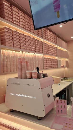A brightly-lit interior with a pink and white color scheme features a wall of stacked pink pizza boxes. A white and pink coffee machine labeled 'Radiofonica' sits on a counter, accompanied by branded pink and white cups, napkins, and dispensers. A digital screen above displays an ice cream cone graphic.