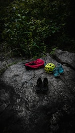 On a large, textured rock surface, several items are neatly arranged, including a red bag, a bright yellow helmet, a pair of blue sneakers, and black shoes. Surrounding the rock, lush greenery with dense leaves adds a natural backdrop.