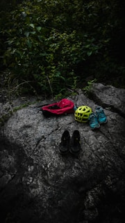 On a large, textured rock surface, several items are neatly arranged, including a red bag, a bright yellow helmet, a pair of blue sneakers, and black shoes. Surrounding the rock, lush greenery with dense leaves adds a natural backdrop.