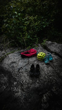 On a large, textured rock surface, several items are neatly arranged, including a red bag, a bright yellow helmet, a pair of blue sneakers, and black shoes. Surrounding the rock, lush greenery with dense leaves adds a natural backdrop.