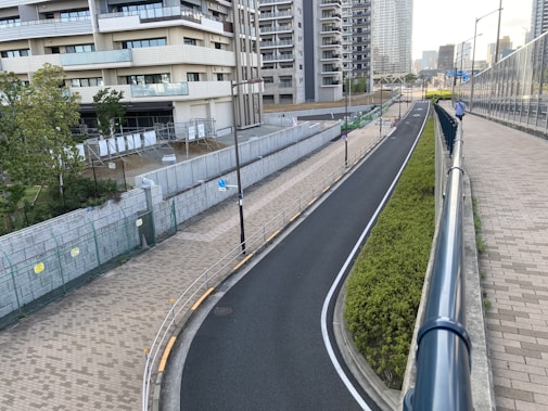 A modern urban scene featuring a winding road flanked by a sidewalk and a green hedge. There are tall residential buildings in the background and a fenced area adjacent to the road. Streetlights line the road, and a clear sky is visible.