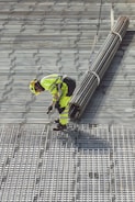 A construction worker in bright safety clothing is standing on a grid-like metal structure. The worker is bending forward, holding onto a bundle of long metal rods suspended by sturdy straps attached to a lifting mechanism.
