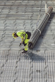 A construction worker in bright safety clothing is standing on a grid-like metal structure. The worker is bending forward, holding onto a bundle of long metal rods suspended by sturdy straps attached to a lifting mechanism.