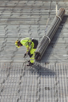 A construction worker in bright safety clothing is standing on a grid-like metal structure. The worker is bending forward, holding onto a bundle of long metal rods suspended by sturdy straps attached to a lifting mechanism.