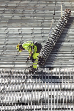 A construction worker in bright safety clothing is standing on a grid-like metal structure. The worker is bending forward, holding onto a bundle of long metal rods suspended by sturdy straps attached to a lifting mechanism.