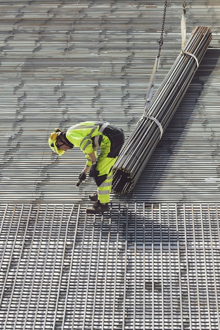 A construction worker in bright safety clothing is standing on a grid-like metal structure. The worker is bending forward, holding onto a bundle of long metal rods suspended by sturdy straps attached to a lifting mechanism.