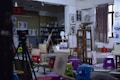 An art studio filled with easels, stools, and various painting supplies. The walls display sketches and busts, creating an atmosphere of creativity and learning. A tripod and several colorful plastic stools are scattered across the room. A large window with a curtain lets in natural light.