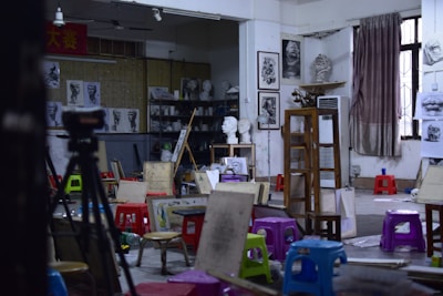 An art studio filled with easels, stools, and various painting supplies. The walls display sketches and busts, creating an atmosphere of creativity and learning. A tripod and several colorful plastic stools are scattered across the room. A large window with a curtain lets in natural light.