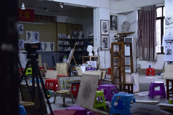 An art studio filled with easels, stools, and various painting supplies. The walls display sketches and busts, creating an atmosphere of creativity and learning. A tripod and several colorful plastic stools are scattered across the room. A large window with a curtain lets in natural light.