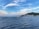Sailboats are calmly anchored in a serene bay with a vibrant blue sea. In the background, a small coastal town is nestled at the foot of a lush, green hill under a sky filled with soft, wispy clouds.