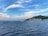 Sailboats are calmly anchored in a serene bay with a vibrant blue sea. In the background, a small coastal town is nestled at the foot of a lush, green hill under a sky filled with soft, wispy clouds.