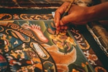 A detailed painting featuring intricate patterns and ornate designs is being meticulously created. The artwork includes mythical creatures, swirling motifs, and vibrant colors. A hand delicately holds a fine paintbrush, applying careful strokes to add detail to the central dragon-like figure.