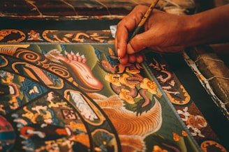 A detailed painting featuring intricate patterns and ornate designs is being meticulously created. The artwork includes mythical creatures, swirling motifs, and vibrant colors. A hand delicately holds a fine paintbrush, applying careful strokes to add detail to the central dragon-like figure.