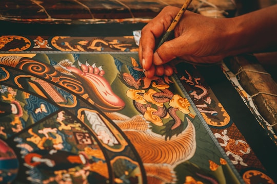 A detailed painting featuring intricate patterns and ornate designs is being meticulously created. The artwork includes mythical creatures, swirling motifs, and vibrant colors. A hand delicately holds a fine paintbrush, applying careful strokes to add detail to the central dragon-like figure.