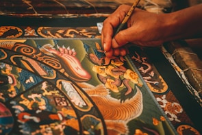 A detailed painting featuring intricate patterns and ornate designs is being meticulously created. The artwork includes mythical creatures, swirling motifs, and vibrant colors. A hand delicately holds a fine paintbrush, applying careful strokes to add detail to the central dragon-like figure.