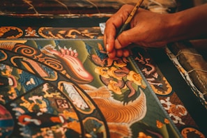 A detailed painting featuring intricate patterns and ornate designs is being meticulously created. The artwork includes mythical creatures, swirling motifs, and vibrant colors. A hand delicately holds a fine paintbrush, applying careful strokes to add detail to the central dragon-like figure.