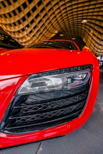A close-up view of the front headlight and grill of a sleek red sports car. The image captures the intricate design details of the car's headlamp and the large black grill with a crisscross pattern. The background features a modern architectural ceiling with a wave-like pattern, enhancing the dynamic feel of the scene.