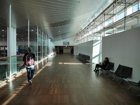 A spacious indoor walkway with wooden flooring and a high ceiling featuring natural light streaming through large windows on the right. A person with a backpack walks along the left side, while another person sits on a chair to the right, using a mobile device. The area appears minimalistic, with rows of empty seating.