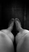 A monochromatic image depicting bare feet and legs stretched out towards a dimly lit, wooden surface. The focus is on the feet, with the legs appearing slightly out of focus, adding depth.