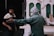 Two individuals are in a workshop-like setting. One person, dressed in a green jacket and grey cap, with a mask, gestures with an outstretched arm, possibly discussing or explaining something. The other person, wearing a two-toned shirt, appears to be listening attentively. The background features partially constructed or decorative panels and tools, suggesting a construction or carpentry environment.