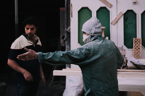 Two individuals are in a workshop-like setting. One person, dressed in a green jacket and grey cap, with a mask, gestures with an outstretched arm, possibly discussing or explaining something. The other person, wearing a two-toned shirt, appears to be listening attentively. The background features partially constructed or decorative panels and tools, suggesting a construction or carpentry environment.