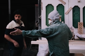 Two individuals are in a workshop-like setting. One person, dressed in a green jacket and grey cap, with a mask, gestures with an outstretched arm, possibly discussing or explaining something. The other person, wearing a two-toned shirt, appears to be listening attentively. The background features partially constructed or decorative panels and tools, suggesting a construction or carpentry environment.