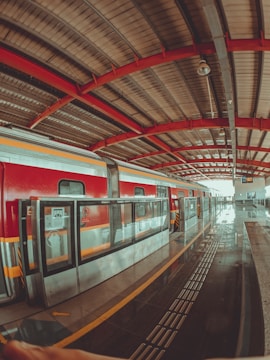 A train station platform with a red and yellow train on the tracks. The platform is covered with a roof, featuring red beams and metal paneling. Safety glass barriers line the edge of the platform alongside tactile paving, indicating where passengers should stand. The area appears clean and well-maintained.