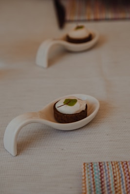 A small dessert sits elegantly on a white ceramic spoon on a textured light-colored tablecloth. The dessert has a dark base and a creamy topping with a small green leaf for decoration. Another similar spoon with dessert is blurred in the background, along with a colorful striped item partially visible at the edge.