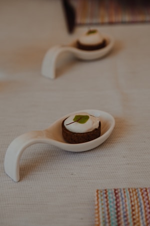 A small dessert sits elegantly on a white ceramic spoon on a textured light-colored tablecloth. The dessert has a dark base and a creamy topping with a small green leaf for decoration. Another similar spoon with dessert is blurred in the background, along with a colorful striped item partially visible at the edge.
